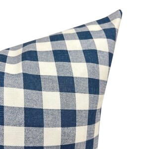 May include: Close-up of a blue and white checkered pillow. The pillow has a classic gingham pattern, with large squares of navy blue and off-white. The fabric appears to be a woven cotton or linen, with a slightly textured surface.