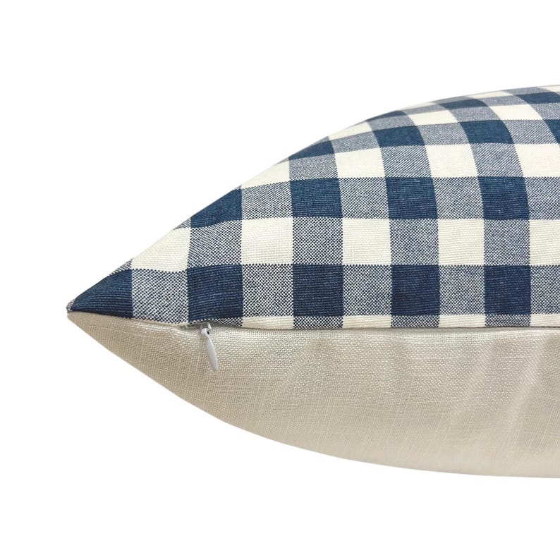 May include: A decorative pillow with a blue and white checkered pattern on the top half and a solid beige linen fabric on the bottom half. A zipper is visible on the side. The pillow is a home decor item.