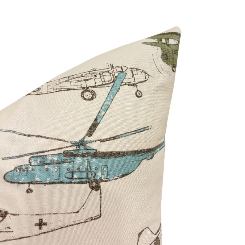 Puede incluir: Coj&iacute;n color crema con un patr&oacute;n repetido de aviones antiguos. El dise&ntilde;o incluye ilustraciones de helic&oacute;pteros y aviones en tonos turquesa, marr&oacute;n y verde oliva. El coj&iacute;n tiene un aspecto suave y texturizado.