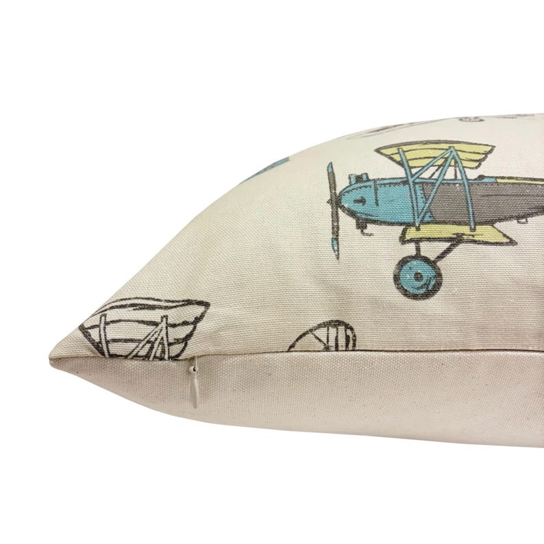 Puede incluir: Coj&iacute;n color crema con un patr&oacute;n repetido de aviones antiguos en azul, amarillo y gris. El coj&iacute;n tambi&eacute;n presenta un barco y otros elementos de dise&ntilde;o. El coj&iacute;n tiene cierre de cremallera.