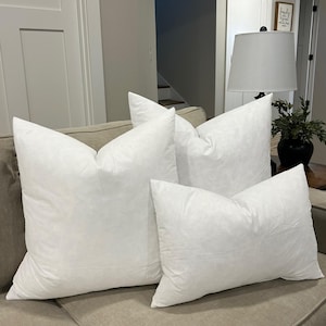 May include: Three white down filled pillow inserts of different sizes on a beige couch. The pillows are arranged in a staggered formation.