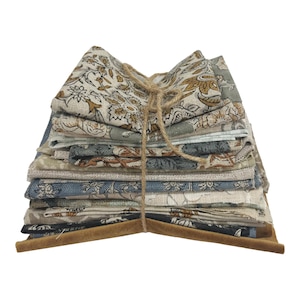 May include: A collection of folded fabric squares featuring floral patterns in beige, blue, and olive green hues. The fabric is bound with twine and sits on a brown, suede-like base. The style is reminiscent of vintage and rustic designs.
