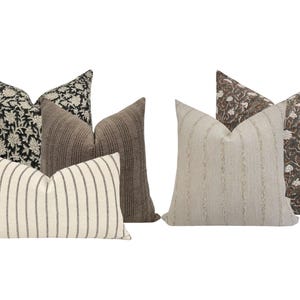 May include: An assortment of decorative throw pillows in various patterns and colors. The pillows include floral, striped, and textured designs in neutral tones of black, white, brown, and beige. The pillows are arranged in a group, showcasing their different styles.