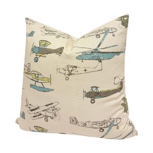 Puede incluir: Coj&iacute;n crema con un estampado repetido de aviones y helic&oacute;pteros antiguos en tonos azules, verdes y marrones. El coj&iacute;n tiene un aspecto suave y acogedor, perfecto para a&ntilde;adir un toque de fantas&iacute;a a cualquier habitaci&oacute;n.
