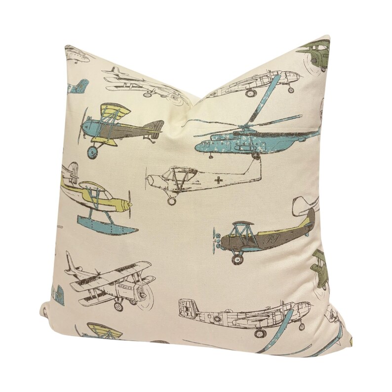Puede incluir: Coj&iacute;n crema con un estampado repetido de aviones y helic&oacute;pteros antiguos en tonos azules, verdes y marrones. El coj&iacute;n tiene un aspecto suave y acogedor, perfecto para a&ntilde;adir un toque de fantas&iacute;a a cualquier habitaci&oacute;n.