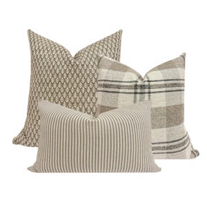 May include: Three decorative throw pillows in neutral tones. One pillow features a repeating leaf pattern, another a plaid design, and the third has vertical stripes. The pillows are in shades of beige, brown, and cream.