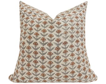 May include: A square throw pillow featuring a repeating floral pattern. The design consists of stylized orange flowers with dark gray stems and leaves printed on a light beige, textured fabric background. The pillow has a plump, filled shape.