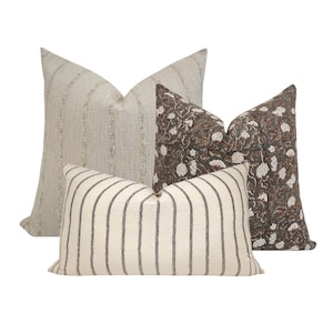May include: Three decorative throw pillows with different patterns. The largest pillow is a light beige with a subtle woven texture. The middle pillow is a white and black striped pattern. The smallest pillow is a dark gray with a brown floral pattern.
