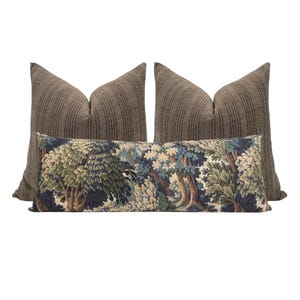 May include: A set of decorative throw pillows. Two large brown textured pillows flank a rectangular pillow with a forest scene in shades of blue, green, and beige. The pillows are arranged against a white background.