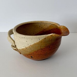 May include: A ceramic pouring bowl featuring a two-tone glaze with a speckled cream upper section and a solid reddish-brown lower section. The bowl includes a small side handle and a distinct pouring spout integrated into the rim.