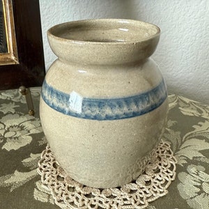 May include: A beige stoneware jar with a blue band, sitting on a cream-colored doily. The jar has a rounded body and a flared rim. The background includes a framed picture and a wooden framed painting.