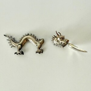 May include: A silver-toned metal dragon figurine separated into two distinct pieces. The left piece is the elongated, arched body of the dragon with textured scales, while the right piece features the detailed dragon head and a small spoon.