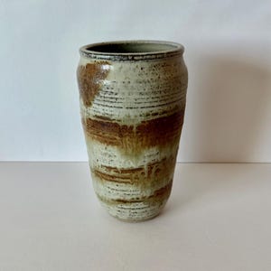 May include: A handmade ceramic vase with a cylindrical shape. The vase features a textured surface with horizontal lines and a color palette of cream, brown, and black. The vase is approximately 8 inches tall.