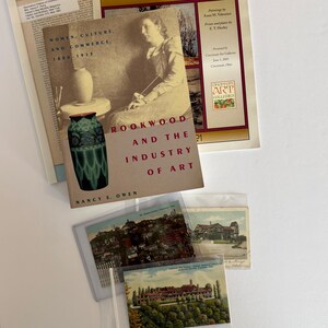 May include: A collection of printed materials including a book titled Rookwood and the Industry of Art, a Beyond Rookwood exhibition catalog, and three vintage postcards featuring landscape views of buildings and cityscapes in Cincinnati, Ohio.