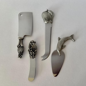 May include: Set of four silver-toned cheese knives and spreaders. Includes a cleaver-style knife, a spreader with a grape cluster design, a serrated knife with an apple-shaped handle, and a spreader with a dolphin design.
