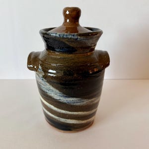 May include: A handmade ceramic jar with a lid. The jar is cylindrical with a brown, black, and white striped pattern. It has two small handles and a brown lid with a small knob. The jar is likely used for storage.