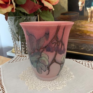 May include: A pink ceramic vase with a floral design featuring a branch with leaves and berries. The vase has a textured surface and a slightly flared rim.