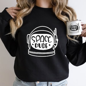 May include: Black sweatshirt with a white astronaut helmet graphic and the text "SPACE DUDE". A white enamel mug with the same design is held in the hand. The design includes stars.