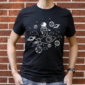 May include: A black t-shirt with a white graphic of an astronaut riding a rocket through space. The graphic includes stars, planets, and clouds.