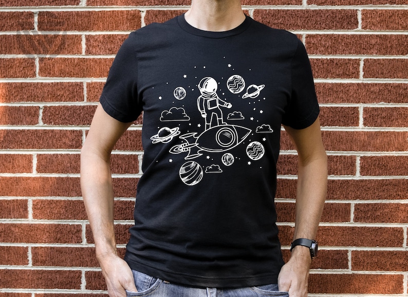 May include: A black t-shirt with a white graphic of an astronaut riding a rocket through space. The graphic includes stars, planets, and clouds.