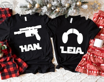 May include: Two black t-shirts with white graphics and text. One shirt features a blaster and the name "HAN", the other features Princess Leia's hair buns and the name "LEIA". Red and black plaid pajama pants and red patterned pajama pants are on either side.