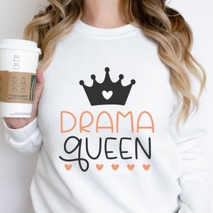 May include: White sweatshirt with a black crown and the text "DRAMA QUEEN" in orange. The crown has a small pink heart in the center. There are five small pink hearts below the text.