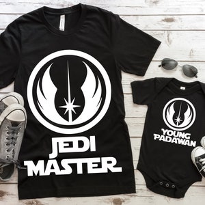 May include: Two black t-shirts with a white Jedi symbol and text. The larger shirt says "Jedi Master" and the smaller shirt says "Young Padawan".