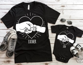 May include: Black t-shirts with white graphic designs of two fists bumping together inside a heart. The larger shirt says "Father" and the smaller shirt says "Son".