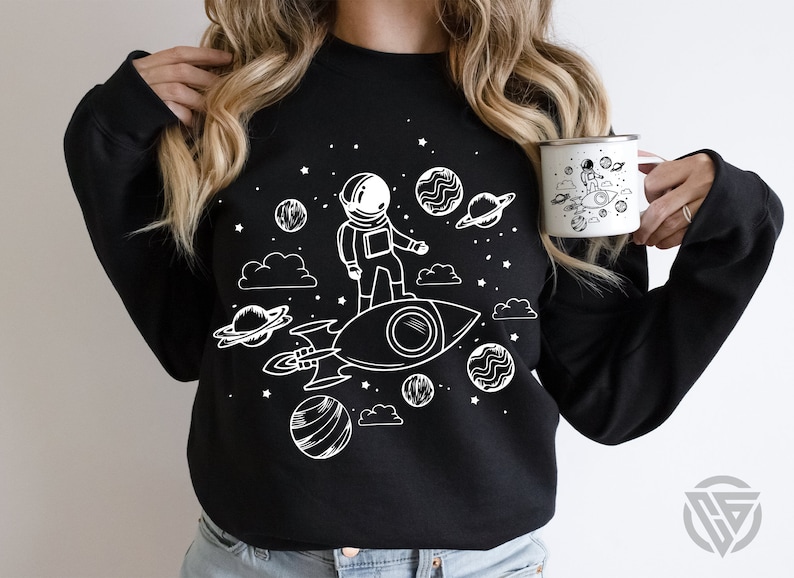 May include: Black sweatshirt with a white graphic of an astronaut riding a rocket through space. The graphic includes stars, planets, and clouds.