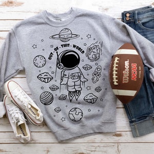 May include: A gray sweatshirt with a black and white illustration of an astronaut in space. The text "OUT OF THIS WORLD" is written around the astronaut. The sweatshirt is laid out on a wooden surface with a football and a pair of white sneakers.