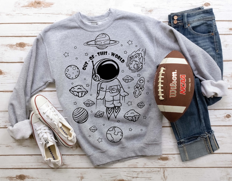 May include: A gray sweatshirt with a black and white illustration of an astronaut in space. The text "OUT OF THIS WORLD" is written around the astronaut. The sweatshirt is laid out on a wooden surface with a football and a pair of white sneakers.