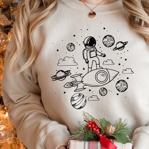 May include: A white sweatshirt with a black and white illustration of an astronaut riding a rocket through space. The illustration includes planets, stars, and clouds.