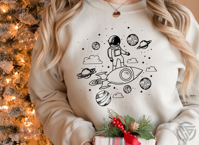 May include: A white sweatshirt with a black and white illustration of an astronaut riding a rocket through space. The illustration includes planets, stars, and clouds.