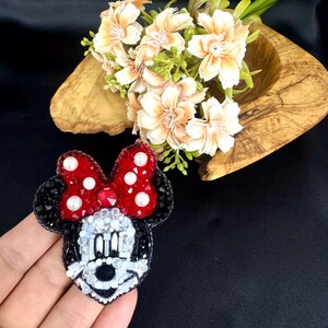 May include: A handcrafted beaded brooch shaped like a cartoon mouse head, featuring a red bow with white pearl accents, black ears, and a white face detailed with clear, black, and white beads. The design is intricate, textured, and colorful.