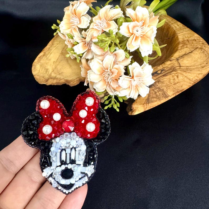 May include: A handcrafted beaded brooch shaped like a cartoon mouse head, featuring a red bow with white pearl accents, black ears, and a white face detailed with clear, black, and white beads. The design is intricate, textured, and colorful.
