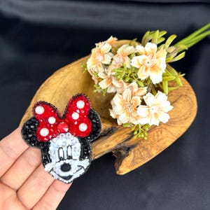 May include: A beaded brooch shaped like a cartoon mouse head with a large red bow featuring white pearl accents. The face is constructed from white, black, and clear beads, while the ears are outlined in black beads with a textured surface.