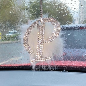 May include: A hanging car ornament featuring a large, silver-toned letter P encrusted with clear, sparkling rhinestones. The letter is layered over a fluffy, white, circular faux fur pom-pom and suspended by a thin, rhinestone-covered chain.