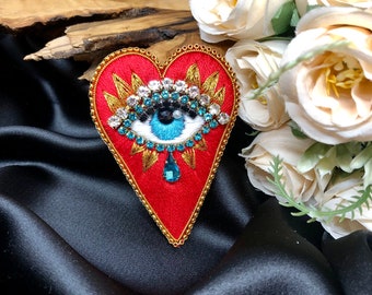 May include: A red heart-shaped embroidered patch with a blue evil eye design surrounded by gold and clear rhinestones. The eye has a white iris and a black pupil with a single teardrop hanging below.