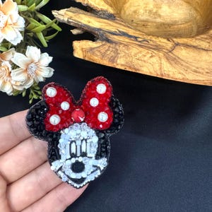 May include: A handcrafted beaded brooch shaped like a cartoon mouse head. The design features a bright red bow with white pearl accents, black ears, and a face detailed with white, clear, and black beads, creating a textured, sparkling appearance.