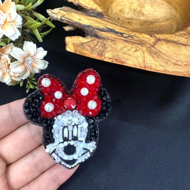 May include: A handcrafted beaded brooch shaped like a cartoon mouse head. The design features a bright red bow with white pearl accents, black ears, and a face detailed with white, clear, and black beads, creating a textured, sparkling appearance.