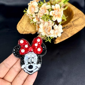 May include: A beaded brooch shaped like a cartoon mouse head with a red bow featuring white pearl accents. The mouse face is crafted with white, black, and clear beads, while the ears are made of black beads. The bow is filled with red crystals.