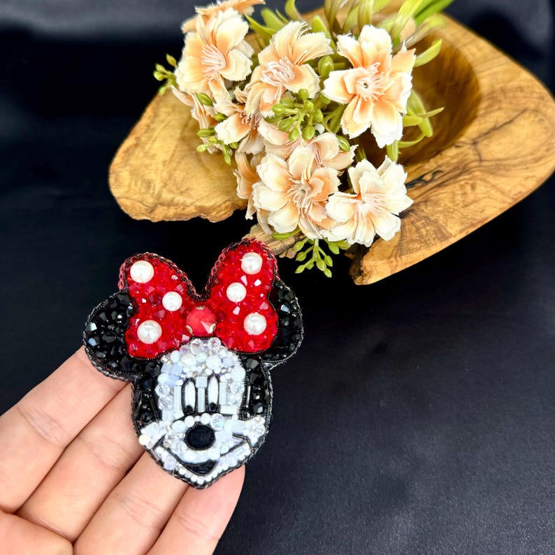 May include: A beaded brooch shaped like a cartoon mouse head with a red bow featuring white pearl accents. The mouse face is crafted with white, black, and clear beads, while the ears are made of black beads. The bow is filled with red crystals.