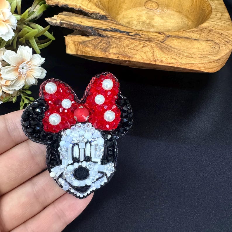 May include: A beaded brooch shaped like a cartoon mouse head with a large red bow featuring white pearl accents. The face is constructed from white, black, and clear beads, while the ears and bow edges are detailed with black and red crystals.