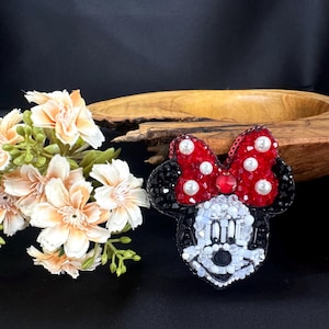 May include: A beaded brooch shaped like a cartoon mouse head with black ears, a red bow with white pearl accents, and a face constructed from white, clear, and black beads. The design features intricate beadwork and sequins throughout the surface.