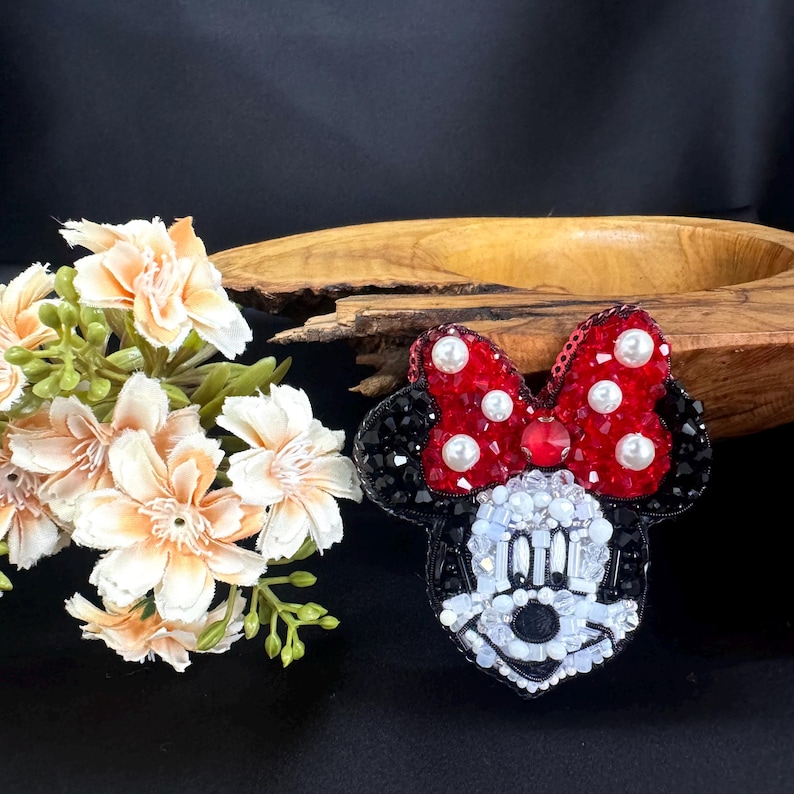 May include: A beaded brooch shaped like a cartoon mouse head with black ears, a red bow with white pearl accents, and a face constructed from white, clear, and black beads. The design features intricate beadwork and sequins throughout the surface.