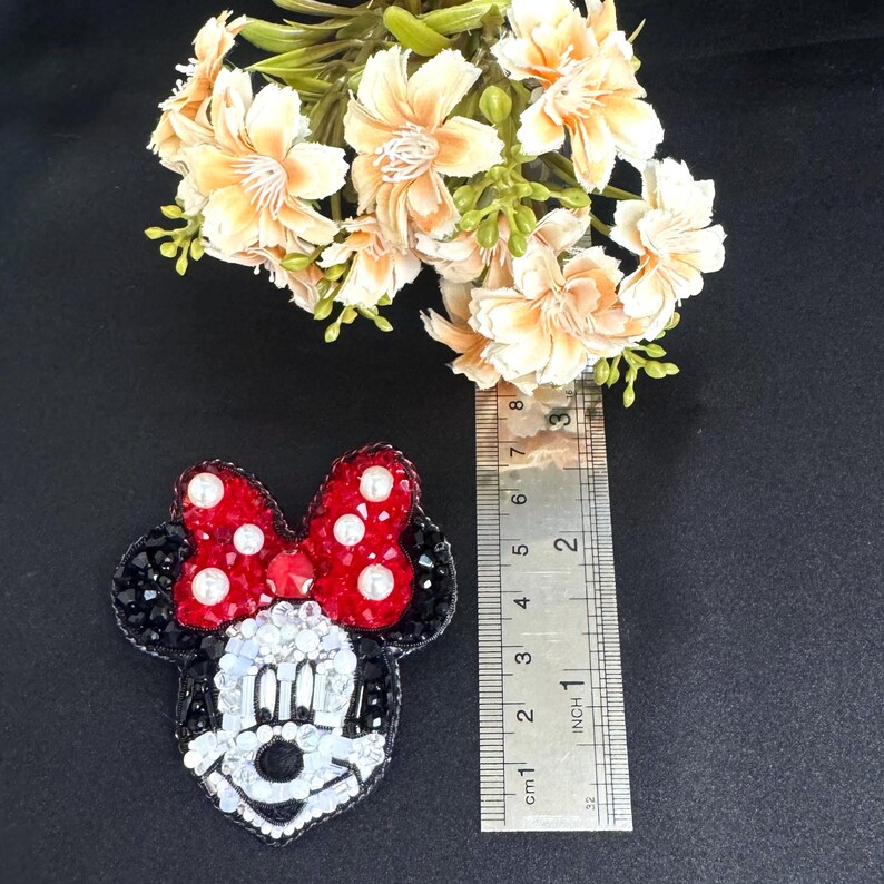 May include: A beaded Minnie Mouse patch featuring a red bow with white pearls, black ears, and a white face with clear crystals. A metal ruler is placed next to the patch, showing it measures approximately 3 inches in height and width.