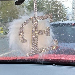 May include: A car rearview mirror hanging ornament featuring a large, capital letter E encrusted with clear, sparkling rhinestones. The letter is attached to a fluffy, white faux fur pom-pom and hangs from a matching rhinestone-covered strap.