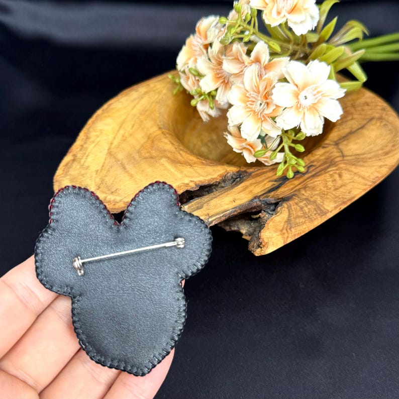 May include: A black, shaped brooch with a silver-toned metal pin mechanism on the back. The item features a dark, stitched border and a smooth, matte-textured surface, designed to be fastened to fabric with its horizontal metal pin closure.