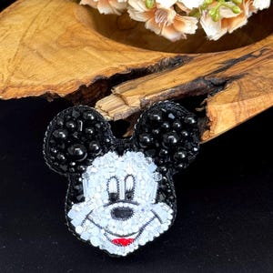 May include: A beaded Mickey Mouse brooch in black and white. The brooch has black beads for the ears and a white and black face with a red mouth. The brooch is set against a dark background.