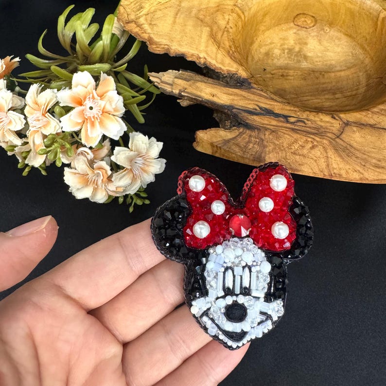 May include: A beaded brooch shaped like a cartoon mouse head with a red bow. The bow features red crystals and white pearl accents. The face is crafted with white, black, and clear beads, including a black nose and defined facial features.